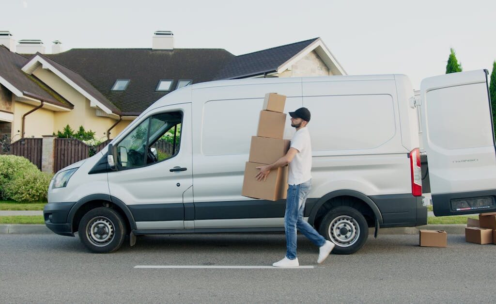 Man unloading cardboard boxes from a delivery van in a suburban neighborhood street.