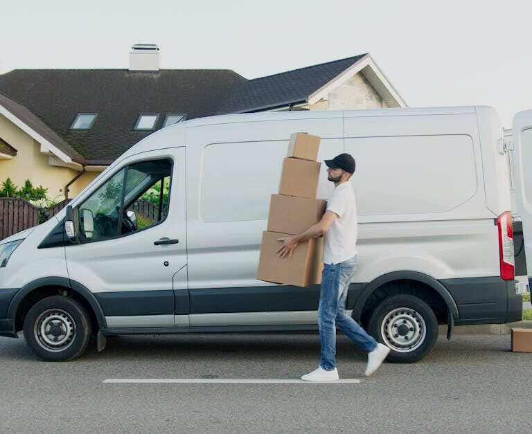 Man unloading cardboard boxes from a delivery van in a suburban neighborhood street.
